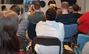 A group of 20 people sitting and looking towards a speaker at a Meetup in a Office-setting.
