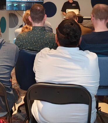 A group of 20 people sitting and looking towards a speaker at a Meetup in a Office-setting.